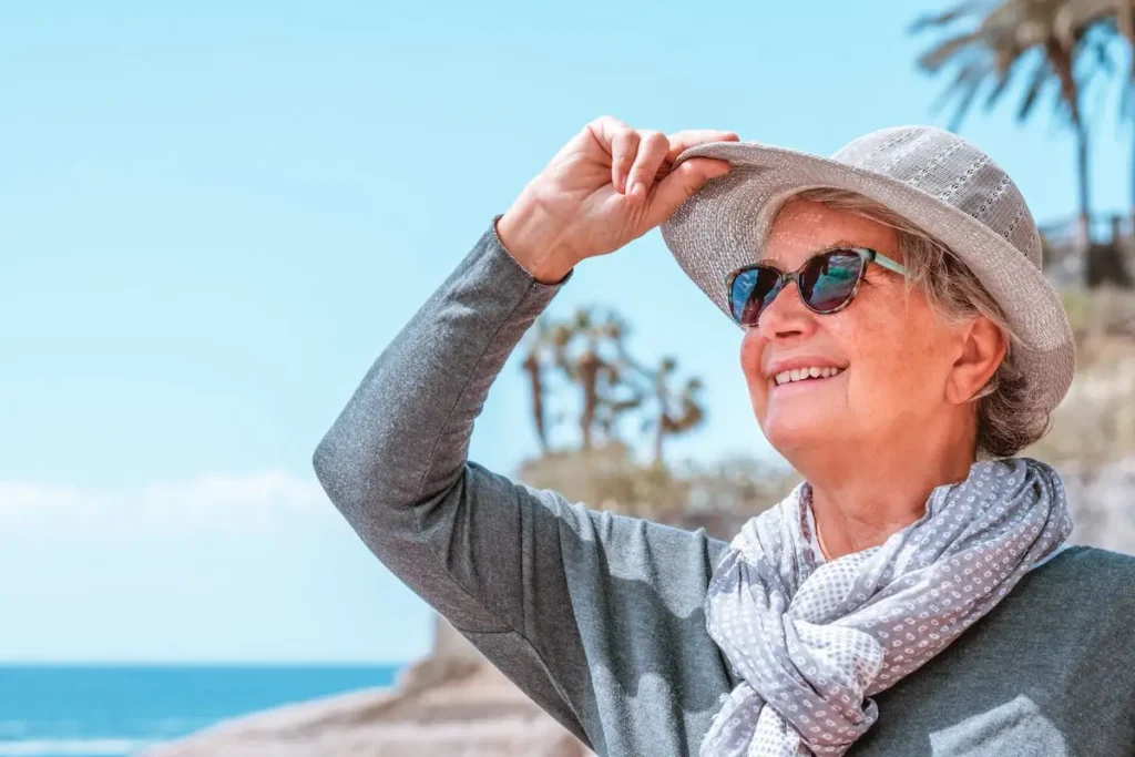 Smiling youthful granny in outdoors at sea enjoying sunny day and vacation, holding her hat looking at horizon over water. Senior carefree woman enjoying retirement and freedom